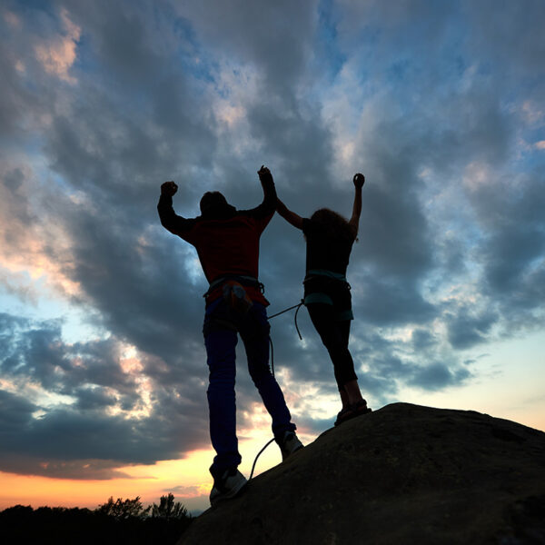 Silhouettes of man and woman on top of mountain. Hikers couple with raised arms on dramatic sky at sunset background.